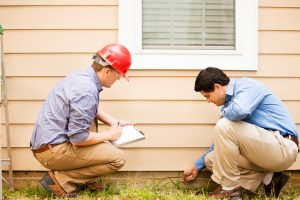 Repairmen, building inspectors, exterminators, engineers, insurance adjusters, or other blue collar workers examine a building/home's exterior wall and foundation. One wears a red hard hat and clear safety glasses and holds a clipboard.  The other checks the foundation with tool.