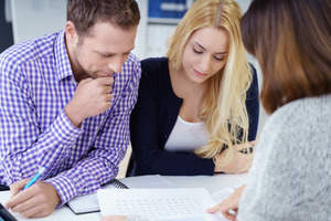 51501459 - broker giving a presentation to a young couple in her office leaning over the desk to explain paperwork to them