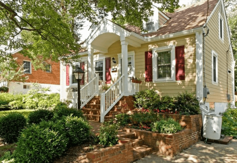 Yellow bungalow with red shutters and lots of bushes out front